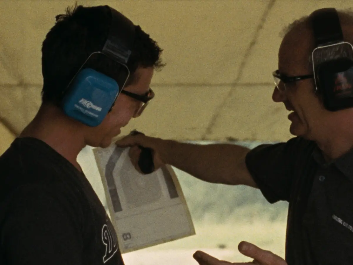 Instrutor mostrando o alvo de papel a um aluno no estande, os dois com protetor auricular